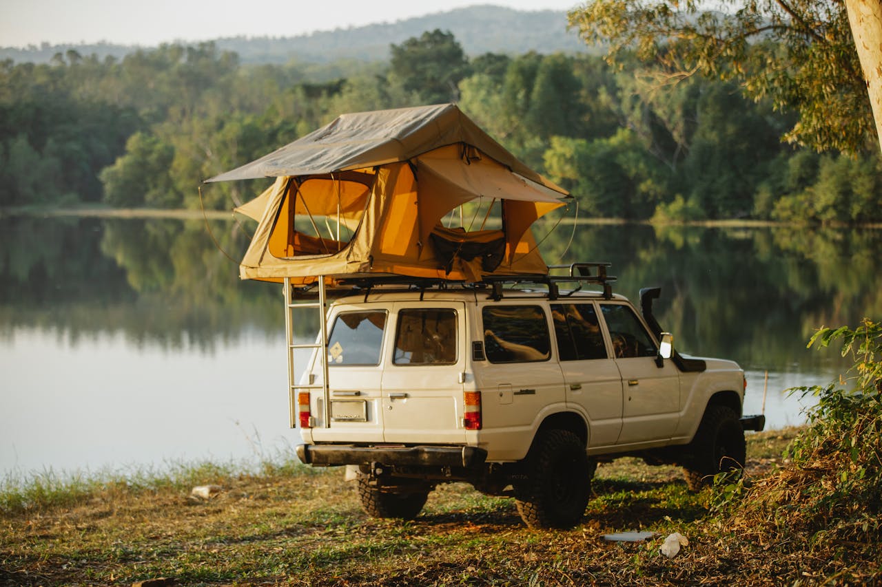 A 4x4 vehicle with a rooftop tent parked by a tranquil lake surrounded by lush forest.